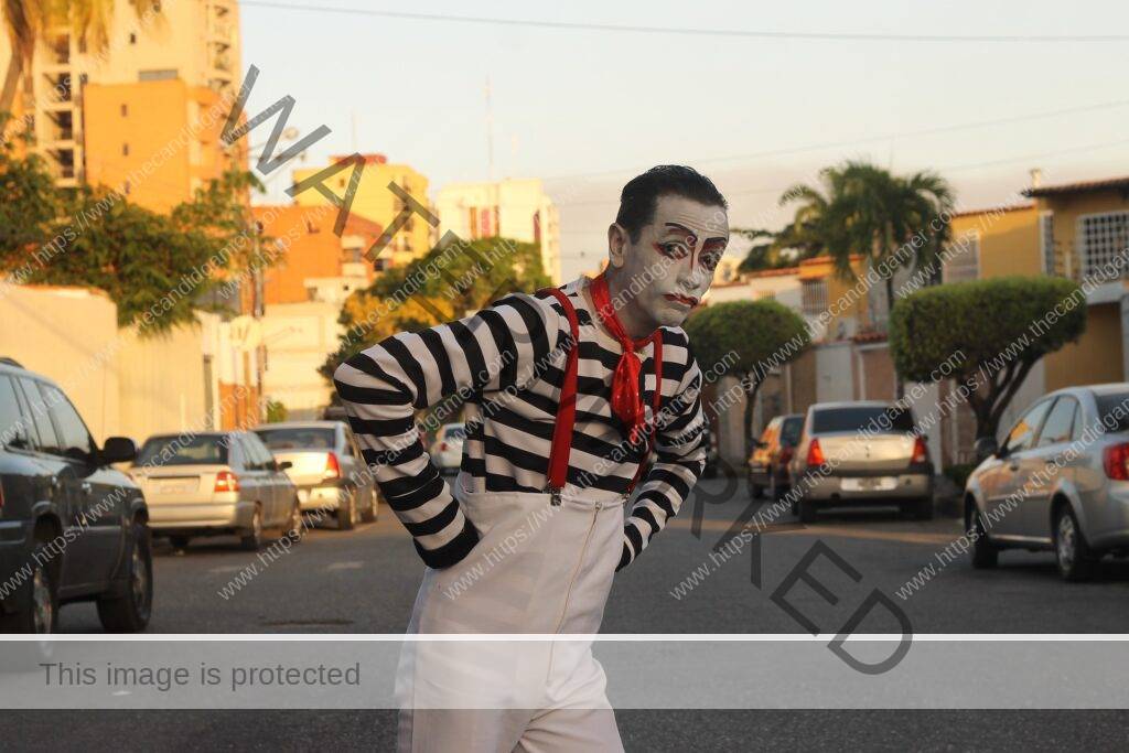 A pantomime stands in a street at midday. His hands are in his pockets, and appears to be shrugging.