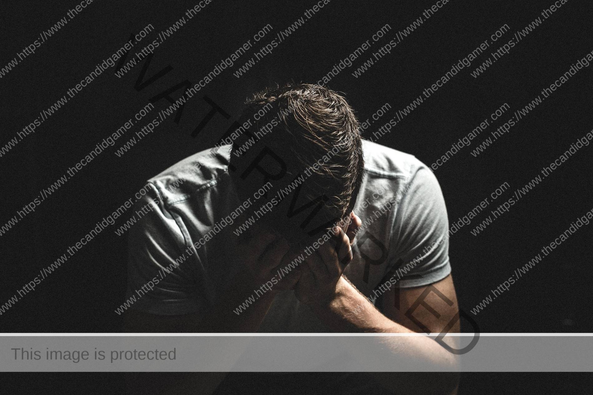 A man sits in the dark, his hands holding his head in an expression of sadness.
