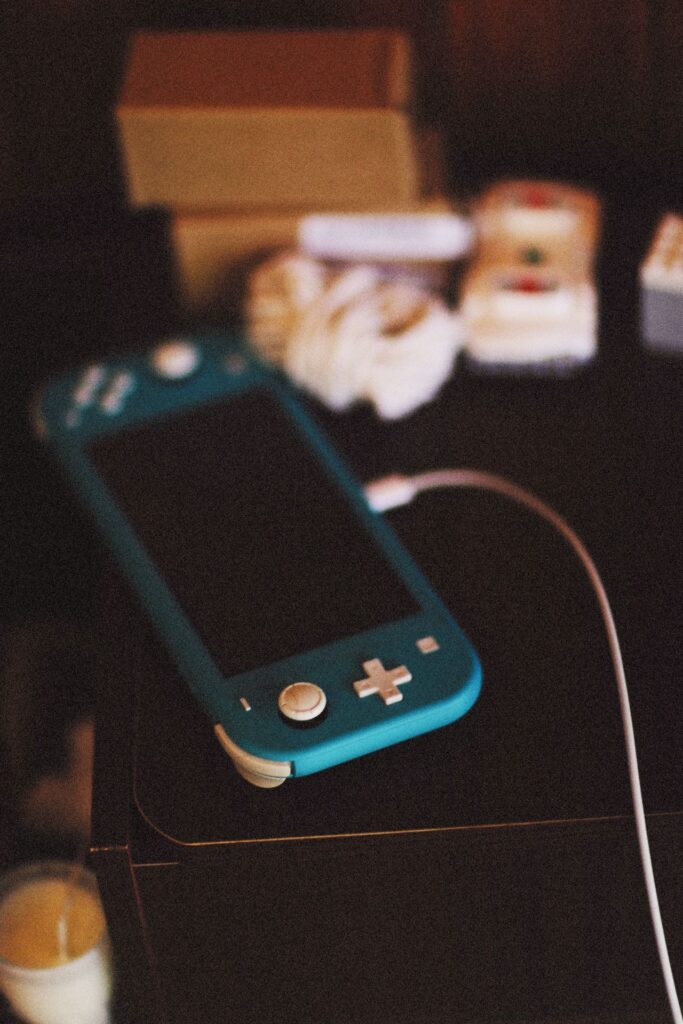 A Blue Nintendo Switch Lite sits on a dark table or nightstand. A charging cord drapes over the table as background objects can be seen out of focus.