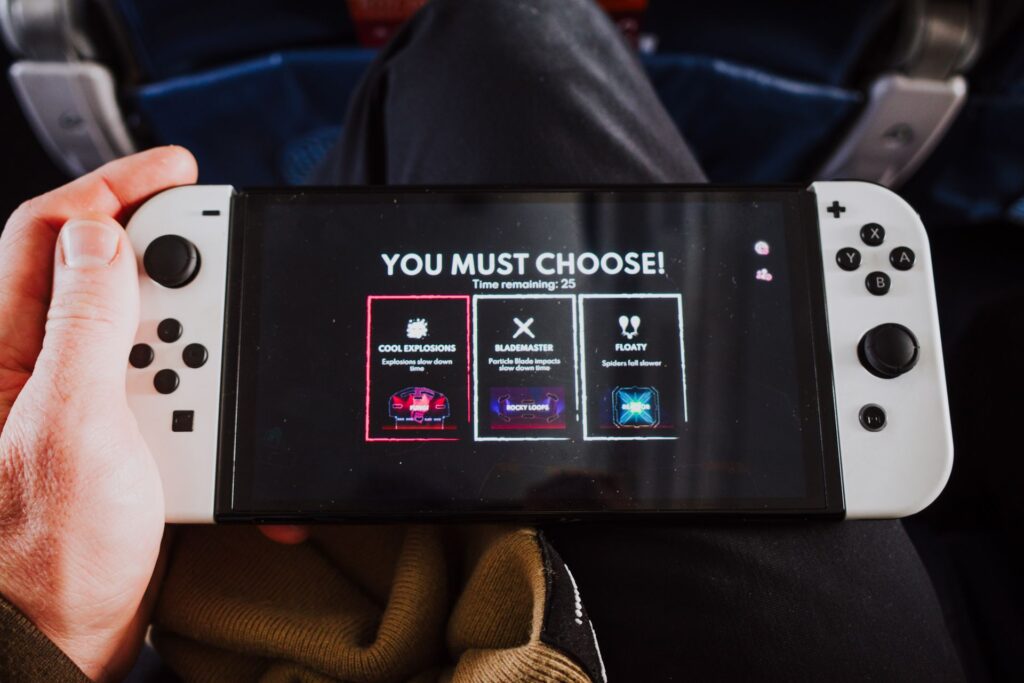 A Switch OLED model being held in handheld mode with one hand, with white Joy-Con connected. The words "You must choose!" can be seen, prompting the player to select a bonus in the game pictured.