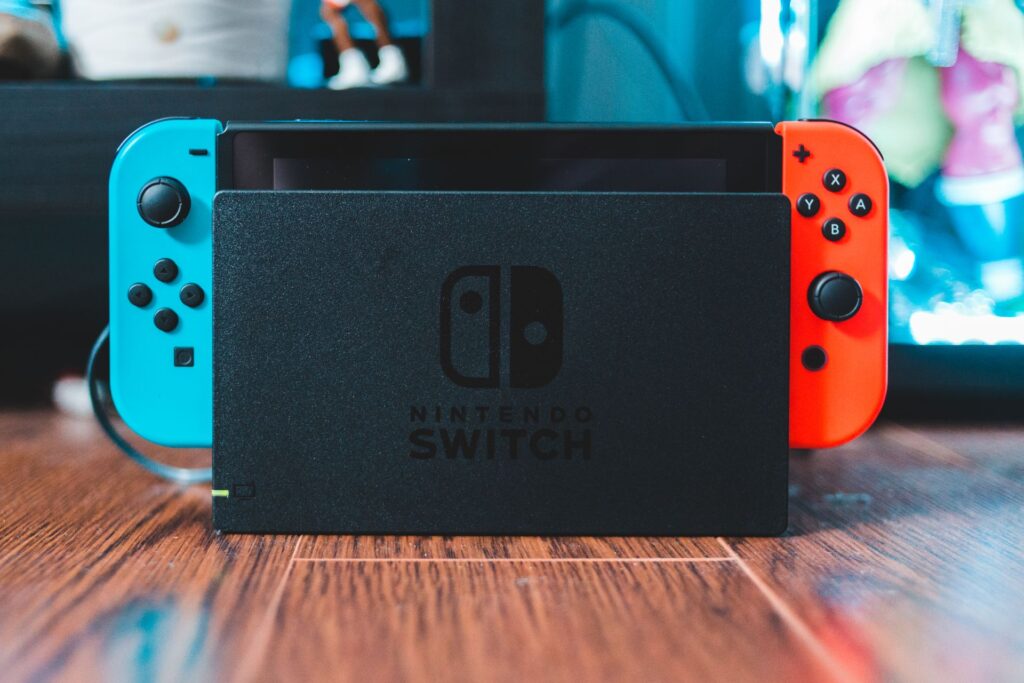 An image of a docked Nintendo Switch on a wooden floor. A window and shelf can be seen out of focus in the background.